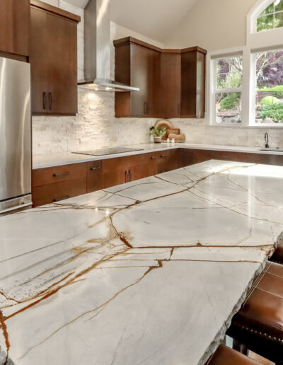 A modern kitchen with wooden cabinets, a large marble island, stainless steel appliances, and a big window overlooking greenery. Three stools are lined up at the island.